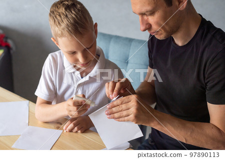 A cute boy is sitting with his dad at the table and collecting origami. Holds a white sheet of paper A cute boy is sitting with his dad at the table and collecting origami. Holds a white sheet of paper 97890113