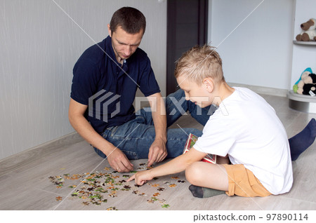 A cute boy with a cheerful dad is collecting a puzzle lying on the floor in a room A cute boy with a cheerful dad is collecting a puzzle lying on the floor in a room 97890114