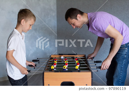 Cute boy playing table football with his emotional dad in the room Cute boy playing table football with his emotional dad in the room 97890119