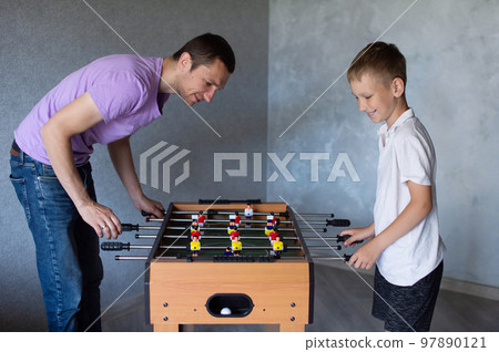 Cute boy playing table football with his emotional dad in the room Cute boy playing table football with his emotional dad in the room 97890121