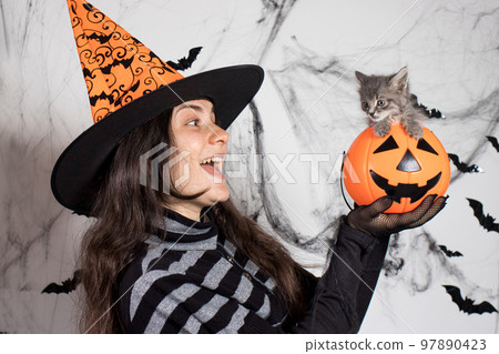 A woman in a witch costume in a hat holds a bucket pumpkin with cat. Halloween people and animal. A woman in a witch costume in a hat holds a bucket pumpkin with cat. Halloween people and animal. 97890423