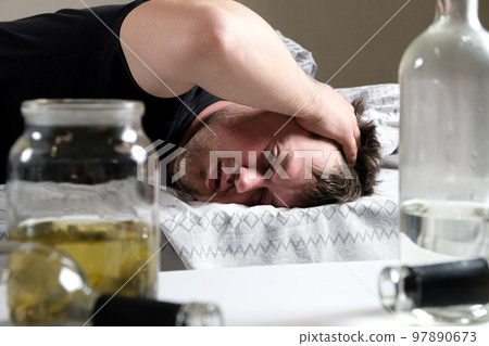 A drunk man lies in bed, holding his hands to his head. Hangover, alcoholic delirium. A drunk man lies in bed, holding his hands to his head. Hangover, alcoholic delirium. 97890673