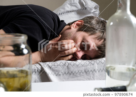 A drunk man lies in bed, holding his hands to his head. Hangover, alcoholic delirium. 97890674