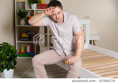 A puzzled, tired father cobbles the baby bed, wipes the sweat off his face. Heavy assembly of home furniture by yourself 97890724