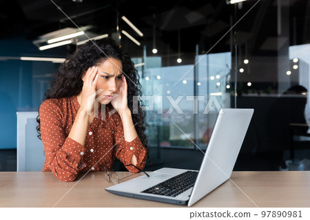 Overtired hispanic business woman working inside modern office, office worker with laptop having severe headache. Overtired hispanic business woman working inside modern office, office worker with laptop having severe headache. 97890981