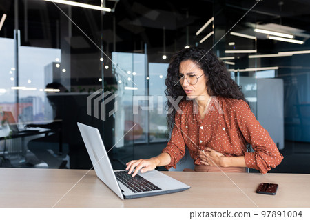 Sick business woman working inside office using laptop while sitting at table, Hispanic woman has severe stomach pain. 97891004