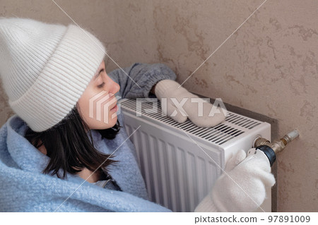 a warmly dressed woman sits by the radiator and checks with her hand whether there is heating a warmly dressed woman sits by the radiator and checks with her hand whether there is heating 97891009