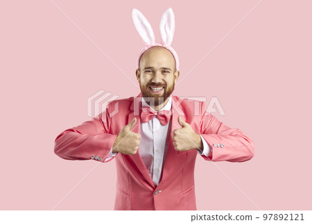 Funny young man in pink suit and Easter bunny ears smiling and showing thumbs up 97892121