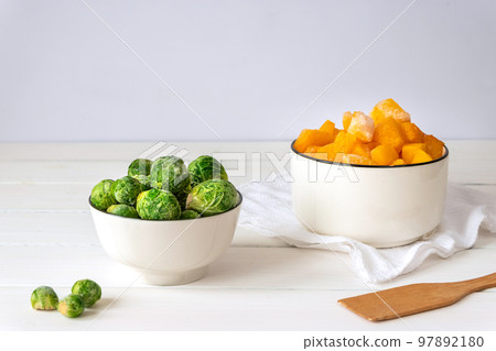 Frozen vegetables assortment. Frozen Brussels sprouts and pieces of pumpkin in white bowls on white table Frozen vegetables assortment. Frozen Brussels sprouts and pieces of pumpkin in white bowls on white table 97892180
