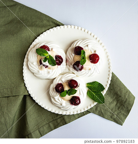 Meringue cakes decorated with whipped cream, cherries and mint leaves on a white plate and green napkin, top view Meringue cakes decorated with whipped cream, cherries and mint leaves on a white plate and green napkin, top view 97892183