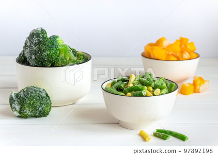 Frozen vegetables assortment. Frozen broccoli, French beans and pumpkin in white bowls on white table Frozen vegetables assortment. Frozen broccoli, French beans and pumpkin in white bowls on white table 97892198