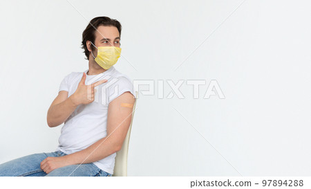 Glad young caucasian male in protective mask with band-aid on shoulder after vaccination shows finger on copy space 97894288