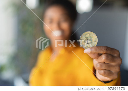 Young black businesswoman showing Bitcoin and smiling, selective focus on golden coin, office interior Young black businesswoman showing Bitcoin and smiling, selective focus on golden coin, office interior 97894373