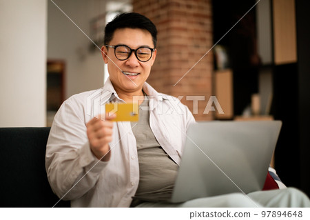 E-commerce concept. Happy asian mature man holding credit card and shopping online with laptop, sitting on couch at home E-commerce concept. Happy asian mature man holding credit card and shopping online with laptop, sitting on couch at home 97894648