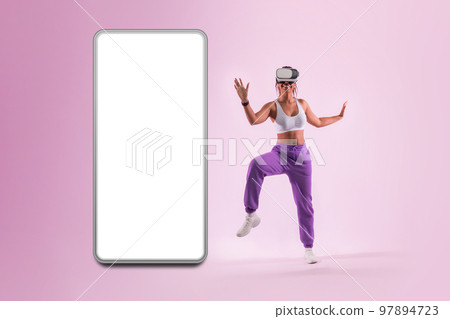 Dance moves in metaverse. Young woman dancing in virtual reality headset near huge smartphone with blank screen Dance moves in metaverse. Young woman dancing in virtual reality headset near huge smartphone with blank screen 97894723