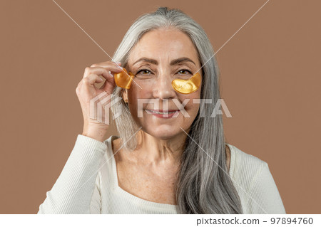 Smiling old caucasian female with natural beauty applies patches under eyes, isolated on brown background, close up Smiling old caucasian female with natural beauty applies patches under eyes, isolated on brown background, close up 97894760