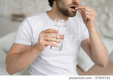 Sad desperate young caucasian guy suffers from pain, illness, takes pills, holds glass of water, sits on bed Sad desperate young caucasian guy suffers from pain, illness, takes pills, holds glass of water, sits on bed 97894782