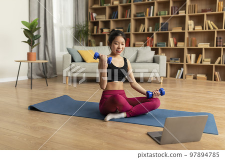 Domestic online sports training. Young asian lady doing exercises with dumbbells in front of laptop, sitting on yoga mat 97894785