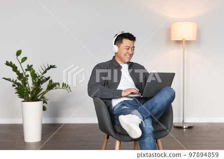 Glad adult chinese guy in wireless headphones listen to music and watch video on laptop in living room 97894859
