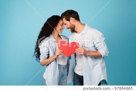 Smiling young european husband in casual and arabic wife hold heart, enjoy tender moment 97894865