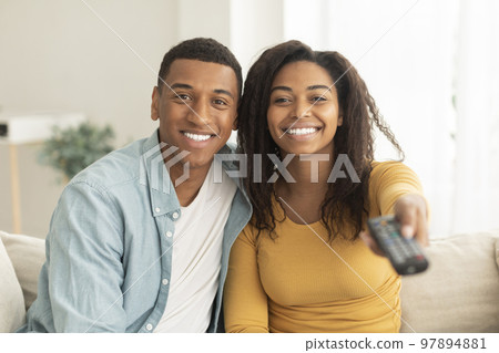 Smiling millennial african american husband hug his wife, switch channel with remote control, watch tv Smiling millennial african american husband hug his wife, switch channel with remote control, watch tv 97894881