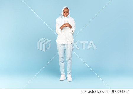 African american woman in hijab using smartphone, texting while standing in studio over blue background, full length 97894895