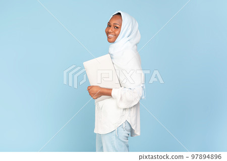 Cheerful black muslim woman holding laptop computer posing over blue studio background, free space 97894896