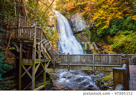 Bridge boardwalk crosses river in front of huge waterfall over cliffs surrounded by fall foliage Bridge boardwalk crosses river in front of huge waterfall over cliffs surrounded by fall foliage 97895152