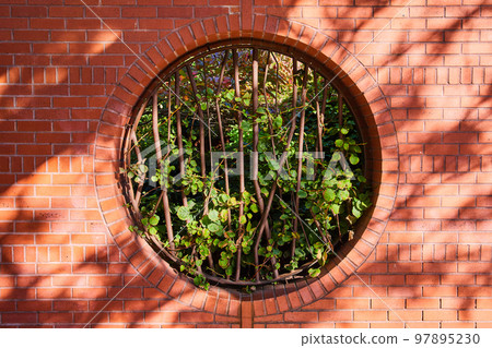 Brick wall with circle opening blocked by abstract steel bars and filled with greenery in center straight on 97895230