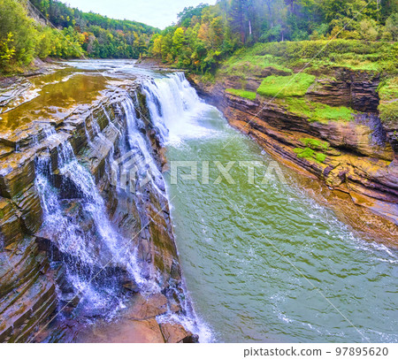 Aerial along stunning waterfall pouring over cliffs with lush forests in background Aerial along stunning waterfall pouring over cliffs with lush forests in background 97895620