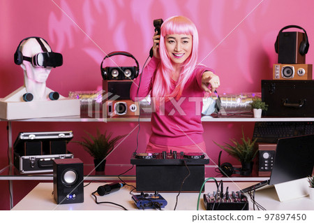 Asian dj listening techno music at headphones while mixing song at professional mixer console, having fun with fans in club at night. Artist with pink hair doing performance with audio equipment 97897450