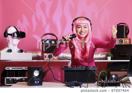 Asian musician sitting at dj table performing techno music using professional turntables, wearing headset and talking with fans using microphone. Artist with pink hair playing electronic song at night 97897464