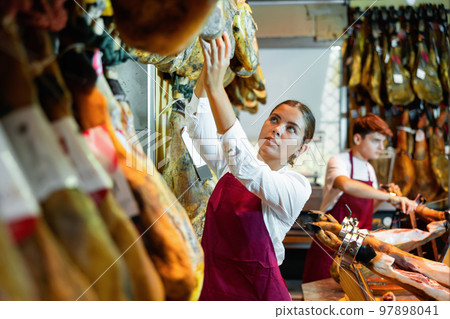 Young salesgirl working at counter of butcher shop selling Iberian jamon Young salesgirl working at counter of butcher shop selling Iberian jamon 97898041