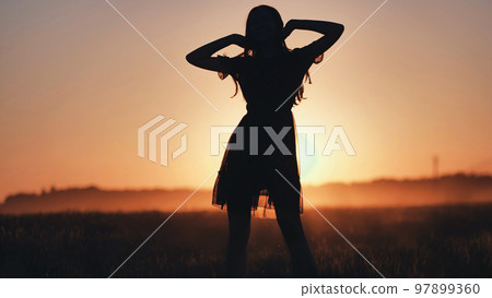 A silhouette of a young girl dancing and spinning on a warm summer evening at sunset. 97899360