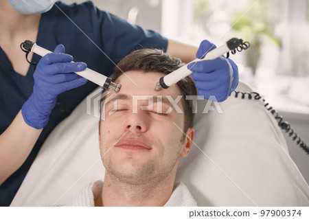Close up of a young man laying in cosmetologist cabinet and has a cavitation procedure for face skin. Female cosmetologist wearing blue medical costume and gloves. Man wearing white bathrobe. Close up of a young man laying in cosmetologist cabinet and has a cavitation procedure for face skin. Female cosmetologist wearing blue medical costume and gloves. Man wearing white bathrobe. 97900374