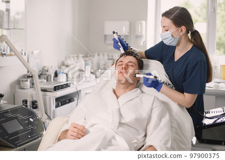 Young man laying in cosmetologist cabinet and has a cavitation procedure for face skin. Female cosmetologist wearing blue medical costume and face mask. Man wearing white bathrobe. Young man laying in cosmetologist cabinet and has a cavitation procedure for face skin. Female cosmetologist wearing blue medical costume and face mask. Man wearing white bathrobe. 97900375