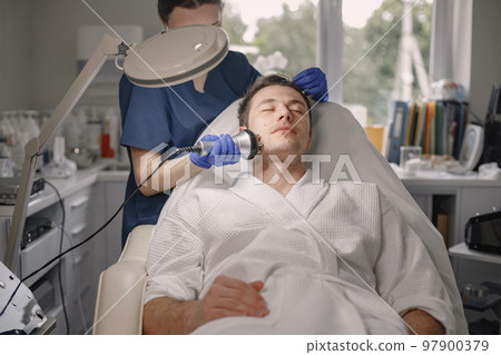 Young man laying in cosmetologist cabinet and has a cavitation procedure for face skin. Female cosmetologist wearing blue medical costume and face mask. Man wearing white bathrobe. Young man laying in cosmetologist cabinet and has a cavitation procedure for face skin. Female cosmetologist wearing blue medical costume and face mask. Man wearing white bathrobe. 97900379