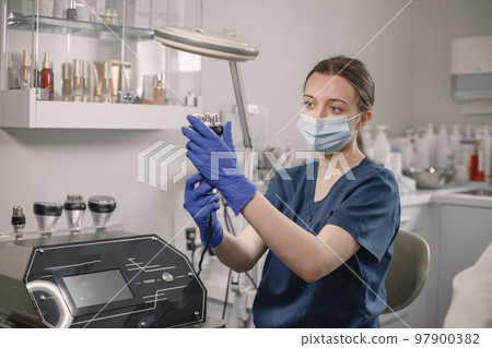 Young female cosmetologist is going to do a cavitation procedure for face skin. Female cosmetologist wearing blue medical costume, gloves and face mask. Woman holding a special equipment. Young female cosmetologist is going to do a cavitation procedure for face skin. Female cosmetologist wearing blue medical costume, gloves and face mask. Woman holding a special equipment. 97900382