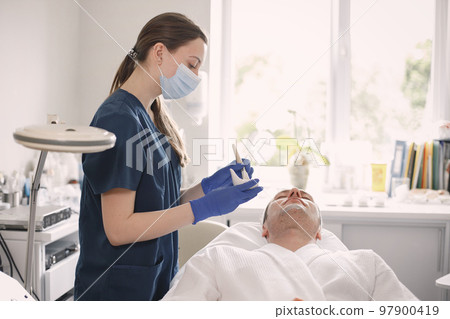 Man laying in cosmetologist cabinet and has a procedure for face skin. Female cosmetologist wearing blue medical costume and face mask. Woman applying a mask on man's face. Man laying in cosmetologist cabinet and has a procedure for face skin. Female cosmetologist wearing blue medical costume and face mask. Woman applying a mask on man's face. 97900419