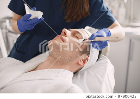 Man laying in cosmetologist cabinet and has a procedure for face skin. Unrecognized female cosmetologist wearing blue medical costume. Woman applying a mask on man's face. Man laying in cosmetologist cabinet and has a procedure for face skin. Unrecognized female cosmetologist wearing blue medical costume. Woman applying a mask on man's face. 97900422