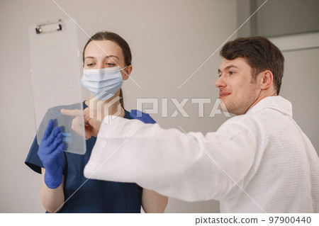 Man sitting in cosmetologist cabinet and talking with cosmetologist. Female cosmetologist wearing blue medical costume and face mask. Man wearing white bathrobe. 97900440