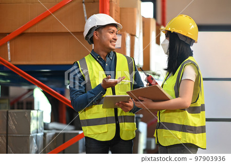 Two warehouse workers in safety uniform working in warehouse full of tall shelves with goods 97903936