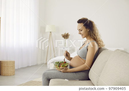 Young pregnant woman sitting on couch and eating bowl of healthy vegetable salad 97909405