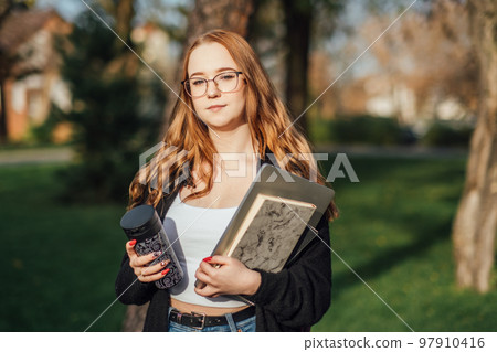 Choosing a university, college. Female college student with books and laptop outdoors. Redhead college student girl with glasses on campus 97910416