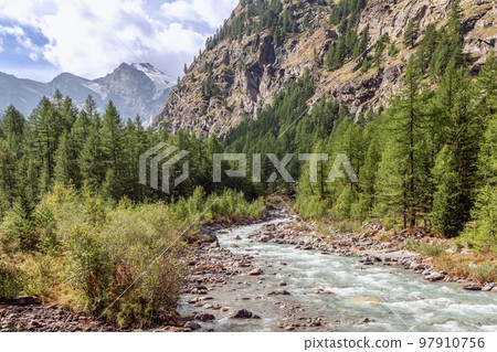 Water stream along rocky rapids in gorge between Italian Alps partly with snow-covered peaks 97910756
