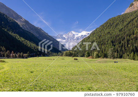 3 trekkers cross flat evergreen famous Alpine Sant Orso meadow surrounded by mountain slopes 3 trekkers cross flat evergreen famous Alpine Sant Orso meadow surrounded by mountain slopes 97910760