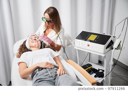 Modern treatment of telangiectasias on skin. Beautician performing local high-precision removal of dilated small vessels on face with innovative yellow laser. 97911266