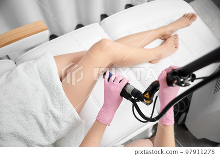 Close up of cosmetologist hands in sterile gloves using erbium ablative laser machine while performing laser treatment on female leg. Young woman having fractional laser procedure in beauty salon. 97911278