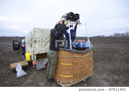Preparations for ballooning: men workers set the gas burner on the top of basket, car and trailer with equipment on a background Preparations for ballooning: men workers set the gas burner on the top of basket, car and trailer with equipment on a background 97911491