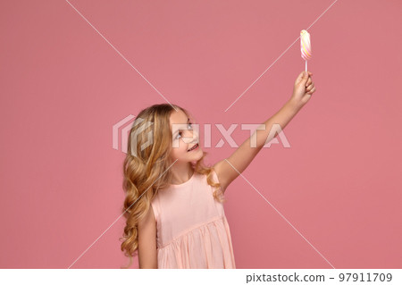 Little girl with a blond curly hair, in a pink dress is posing with a candy 97911709